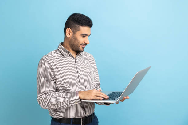 istock Smiling businessman standing with portable computer in hand, looking at display, typing on keyboard. 1436259489