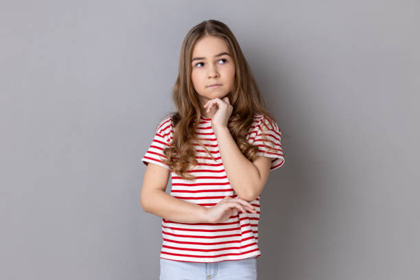 pensive little girl wearing striped t-shirt standing holding chin, thinks about something important. - pensive question mark teenager adversity imagens e fotografias de stock