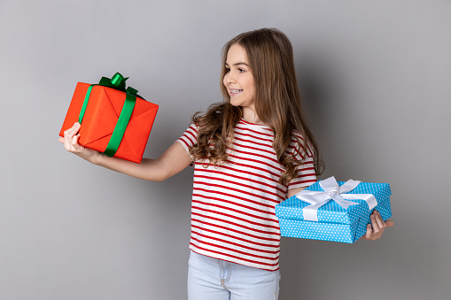 Portrait of charming little girl wearing striped T-shirt celebrating her birthday, holding two present boxes in hands, being happy with gifts. Indoor studio shot isolated on gray background.