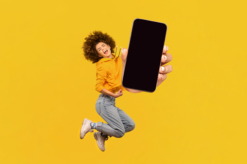 Portrait of extremely happy joyful woman with Afro hairstyle jumping in the air with smart phone in hands, showing empty display for promotion. Indoor studio shot isolated on yellow background.