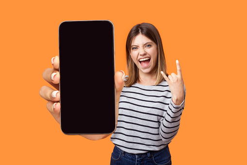 Portrait of excited woman wearing striped shirt standing showing cell phone with empty display, showing rock and roll gesture, screaming happily. Indoor studio shot isolated on orange background.