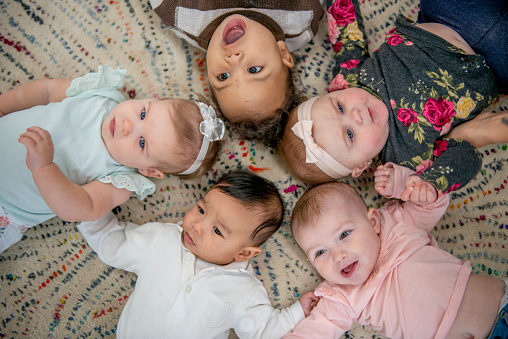 Five babies lay on the floor in a circle with their heads close together as they pose for a portrait.  They are each dressed casually and are smiling in this aerial view photo.