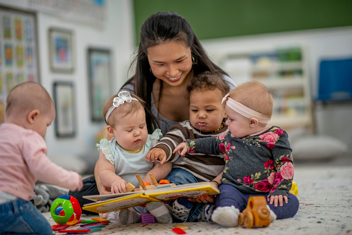A female daycare worker, of Asian decent, sits on the floor with a small group of babies as she supervises them in the Childcare Centre.  She has the babies in close to her as she holds out some puzzles and they play with various toys around them.