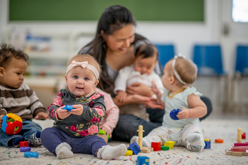 A female Childcare provider sits n the floor with a small group of babies around her.  She is holding one baby on her lap while she supervises the other children playing on the rug in front of her.