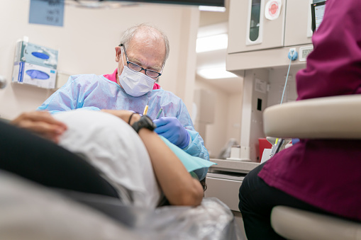 A sixty-five year old dentist doing oral care on an ethnic pregnant patient. He is wearing scrubs and surgical gloves as he meticulously cares for her teeth.