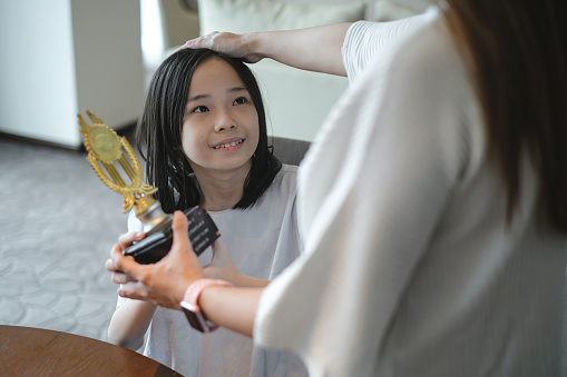 Shot of an unrecognizable Asian mother praising her smiling teenage daughter for winning a school competition. She's holding the trophy in one hand and caressing her daughter's head with the other hand.