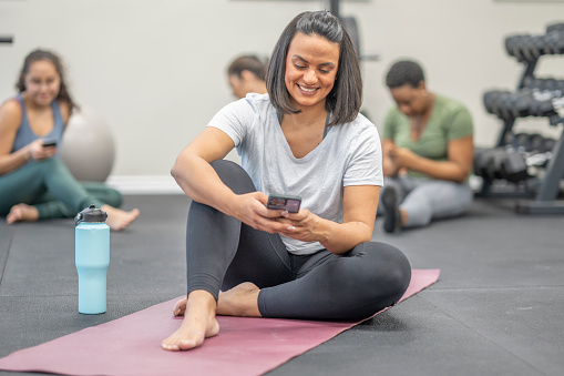 A middle aged woman of Hispanic decent, sits on a Yoga mat at the gym as she  holds her cell phone in her hands and scrolls through.  She is dressed comfortably in athletic wear as she takes a moment to catch up on social media.