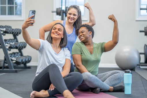 Three middle aged woman sit side-by-side at the gym flexing their muscles as one woman holds out her cell phone to take a picture.  They are each dressed comfortably in athletic wear as they take a break to chat, hydrate and catch up socially with their phones.