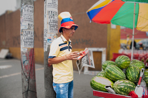 Bogota colombia 8 de Octubre 2015\nMan with hat and colors of the Colombian flag reading a newspaper and selling watermelons