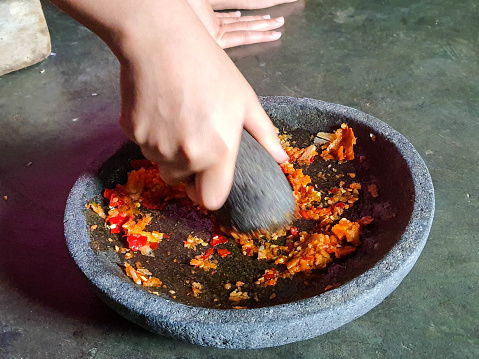 The process of making a chili sauce. A hand is pulverizing onions and chilies in a pestle on a stone mortar to make a chili sauce.