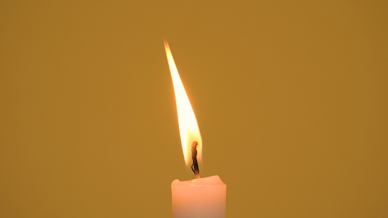 Close-up of a single wax candle burning. Isolated glowing candle against a yellow background. Background or illustration of religious ceremony, spiritual memory, faith and traditions. Camera zoom out.