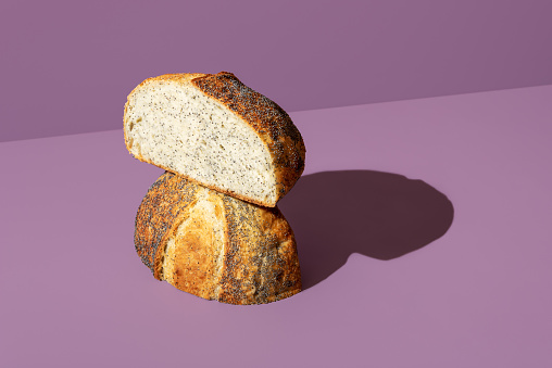 Freshly-baked bread with poppy seeds minimalist on a purple table. Homemade loaf of bread sliced in half in bright light against a colorful background.