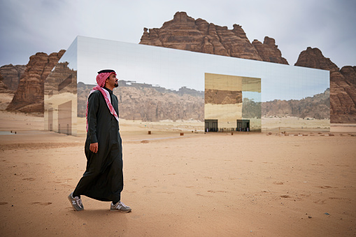 Full length view of man in traditional clothing walking in desert surrounding renowned entertainment venue and largest mirrored building in the world.