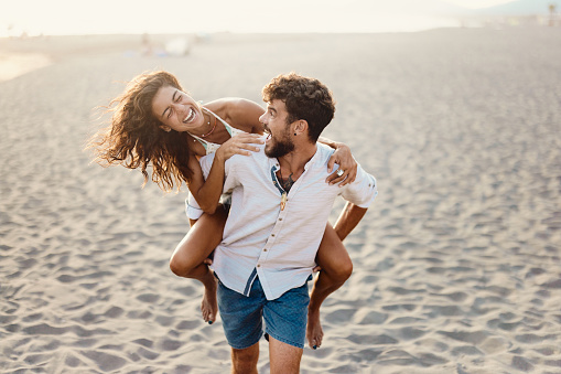 Young playful couple having fun while piggybacking on the beach. Copy space.