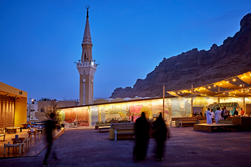 Focus on background group of people talking under illuminated bazaar canopy with minaret and sandstone mountains in background beneath clear blue sky.