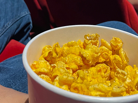 Stock photo of bucket of popcorn in cinema / movie theatre, movie snack. Caramelised / caramel toffee popcorn as movie snack food in paper cups with empty red seats in foreground.