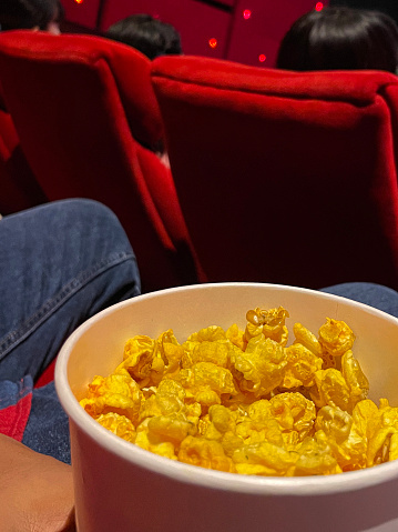 Stock photo of bucket of popcorn in cinema / movie theatre, movie snack. Caramelised / caramel toffee popcorn as movie snack food in paper cups with empty red seats in foreground.
