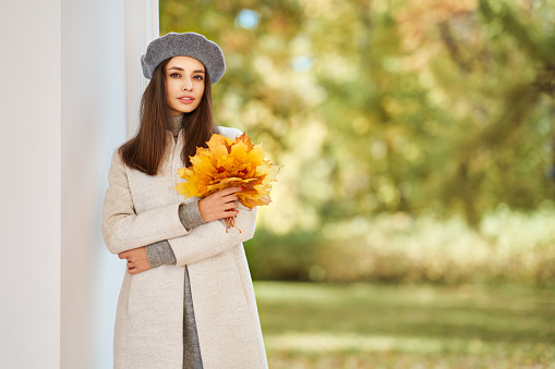 Autumn portrait of a beautiful happy smiling woman with yellow maple leaves in a gray beret in the park
