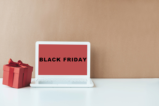 White laptop on white table  with red gift box in front of white wall with brown colored diagonal part. Black Friday text on device screen.