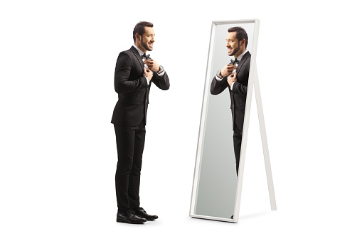 Full length profile shot of a young elegant man getting ready in front of a mirror isolated on white background