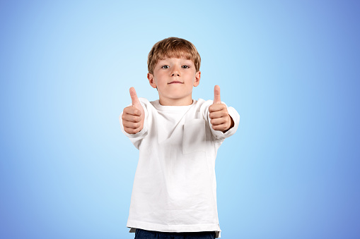 Child boy looking at the camera and showing thumbs up, copy space empty blue background. Concept of like and approval