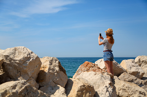 Summer dressed young woman taking photos on vacations with her phone. Standing on the beach of rocks.