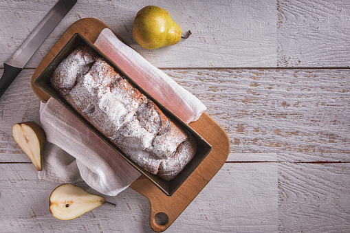 Sweet white bread with powdered sugar and pear lying on a wooden board with a towel and a knife on the table