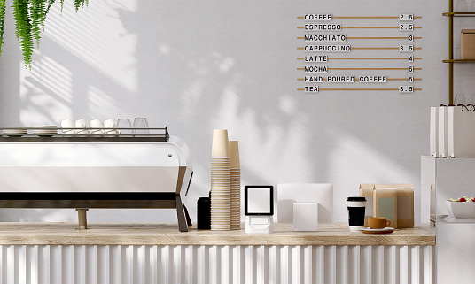 Modern and luxury design of cafe counter with wooden top, espresso machine, digital tablet computer cash register, stack of paper coffee cups with sunlight and leaf shadow from window on white wall