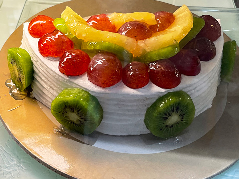 Stock photo showing close-up view of bakery display cabinet of an indulgent dessert of half a fruit and cream gateau, decorated with sliced kiwi, pineapple and red grapes, on circular cake board available for purchasing by the slice.