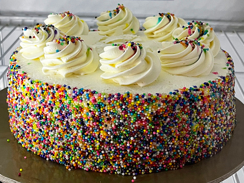 Stock photo showing close-up view of bakery display cabinet of an indulgent rainbow cake dessert, decorated with sugar sprinkles and piped, buttercream swirls, on circular cake board available for purchasing by the slice.