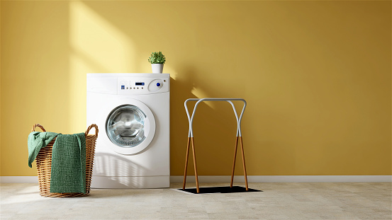 White modern washing machine, rattan basket with towel and cloth rack on tile floor in empty vibrant yellow wall laundry room with beautiful sunlight from window for washing and cleaning product display