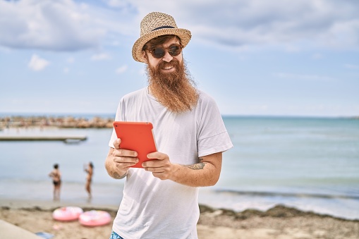 Young redhead tourist man smiling happy using touchpad at the beach.