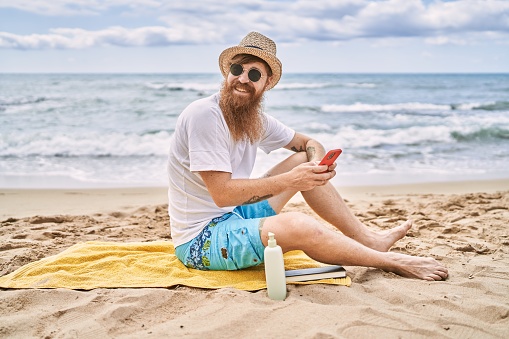 Young redhead man using smartphone sitting on the towel at the beach.