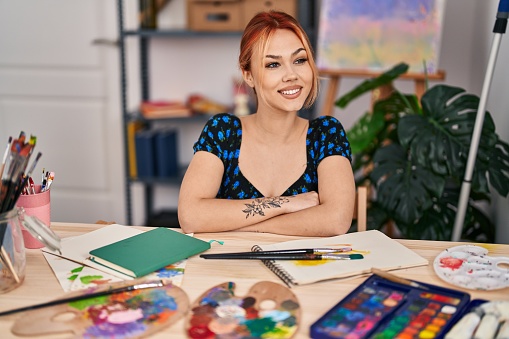 Young caucasian woman artist smiling confident sitting with arms crossed gesture at art studio