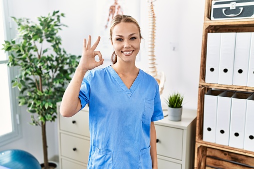 Young caucasian woman working at pain recovery clinic smiling positive doing ok sign with hand and fingers. successful expression.
