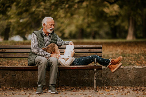 Handsome grandfather spending time with his granddaughter on bench in park on autumn day