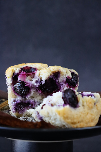 Stock photo showing close-up view of black cake stand containing an individual blueberry muffin unwrapped from a brown paper cake case, against a mottled grey background. Home baking concept.