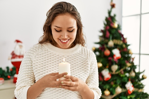 Young beautiful hispanic woman wearing holding candle standing by christmas tree at home