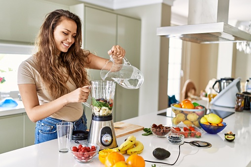 Woman holding blender and making healthy smoothie with various vegetables and fruits in the kitchen at home