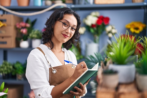 Young beautiful hispanic woman florist smiling confident writing on notebook at flower shop