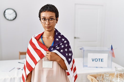 Young hispanic woman with short hair at political campaign election holding usa flag thinking attitude and sober expression looking self confident