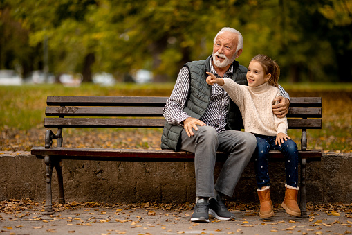 Handsome grandfather spending time with his granddaughter on bench in park on autumn day