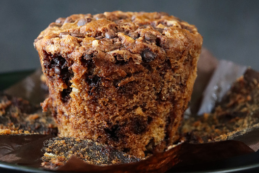 Stock photo showing close-up view of black cake stand containing an individual chocolate chip muffin unwrapped from a brown paper cake case against a mottled grey background. Home baking concept.