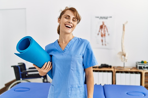 Young caucasian physio therapist girl smiling happy holding yoga mat at the clinic.