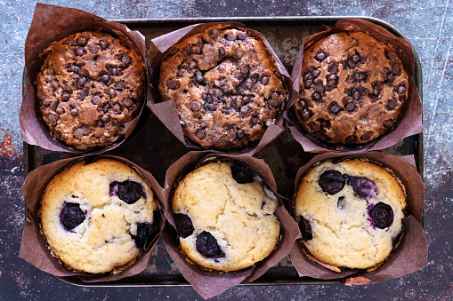 Stock photo showing elevated view of muffin baking tray with baked on dirt containing homemade blueberry and chocolate chip muffins in brown paper cake cases on a mottled blue and brown surface. Home baking concept.