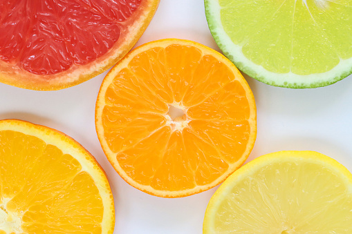 Stock photo showing citrus fruit slices on white background, modern minimalist photo of circular sliced pink grapefruit, oranges, lemon and lime citrus fruits showing segments, seeds / pips and rind around edge with leaves, healthy eating poster wallpaper background design.