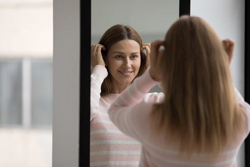 Young beautiful woman in casual sweater, preening, do morning routine, touch her hair standing in front of mirror smiling admire herself feels satisfied with appearance and complexion. Beauty, fashion