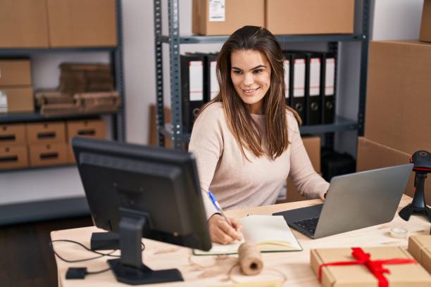 istock Young beautiful hispanic woman ecommerce business worker using laptop writing on notebook at office 1436378090