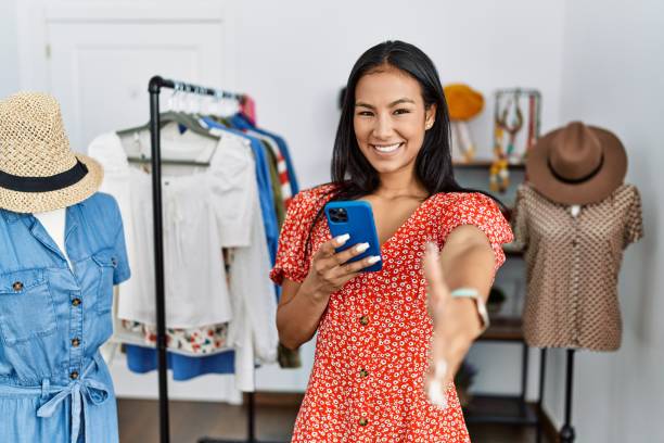 istock Young hispanic woman working at retail boutique using smartphone smiling friendly offering handshake as greeting and welcoming. successful business. 1436378264