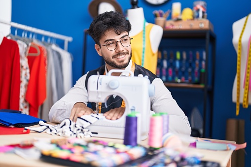 Young hispanic man tailor smiling confident using sewing machine at sewing studio